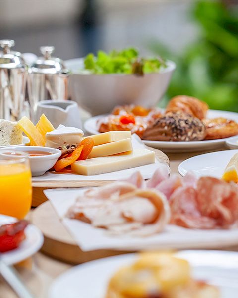 The image shows a breakfast spread with various dishes including cheese, cold cuts, bread, pastries, fruit, and orange juice, arranged on a table.