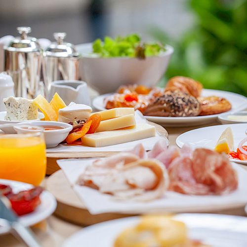 The image shows a breakfast spread with various dishes including cheese, cold cuts, bread, pastries, fruit, and orange juice, arranged on a table.