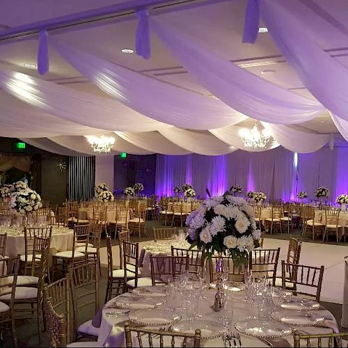 A decorated event hall with round tables, elegant chairs, floral centerpieces, and draped ceilings with purple uplighting and chandeliers.