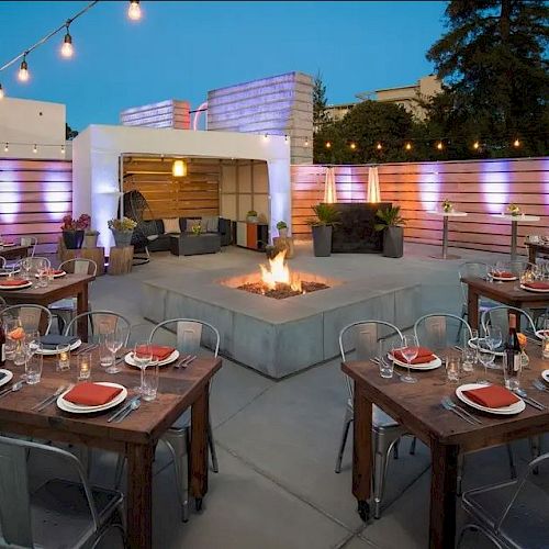 An outdoor dining area with string lights, a fire pit, wooden tables set with red napkins, and a seating area in the background.