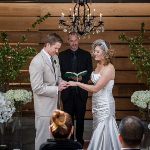 A couple is exchanging rings at their wedding ceremony, officiated by a man holding a book. Decor includes white flowers and green branches.