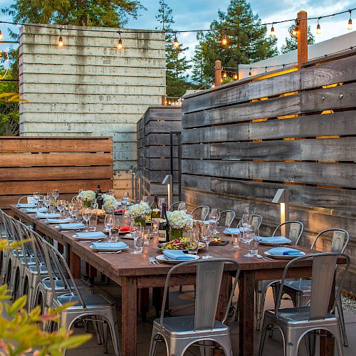 An outdoor dining setup with a long wooden table, metal chairs, and table settings, surrounded by wooden fences and string lights above.