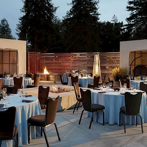 The image depicts an outdoor dining setup with round tables covered in blue tablecloths, chairs, and ambient lighting near a pool surrounded by trees.