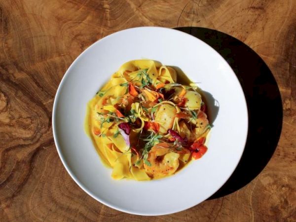 A plate of pasta with shrimp, garnished with herbs and vegetables, is set on a wooden table.