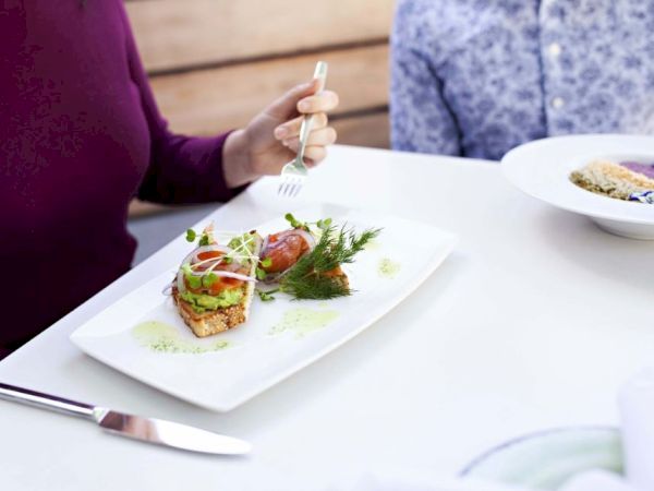 A person holding a fork is about to eat a well-presented dish featuring vegetables and greens on a white plate. Another person is seated beside.