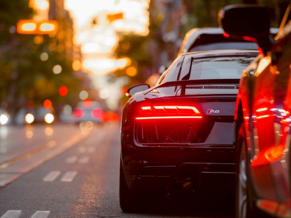 A sleek sports car with illuminated tail lights is parked on a city street at sunset, with blurred lights and buildings in the background.
