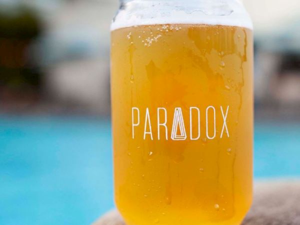 A glass with the word "Paradox" printed on it, filled with light-colored beer, is placed on a wooden surface near a pool.