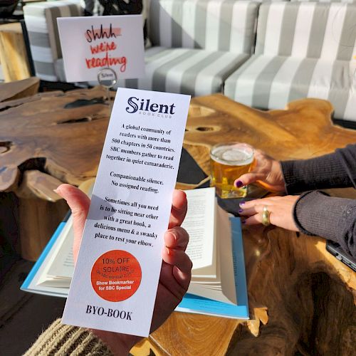 The image shows a person holding a bookmark that reads "Silent" near another person with a drink, in a cozy setting with a sign "Shhh, we're reading".