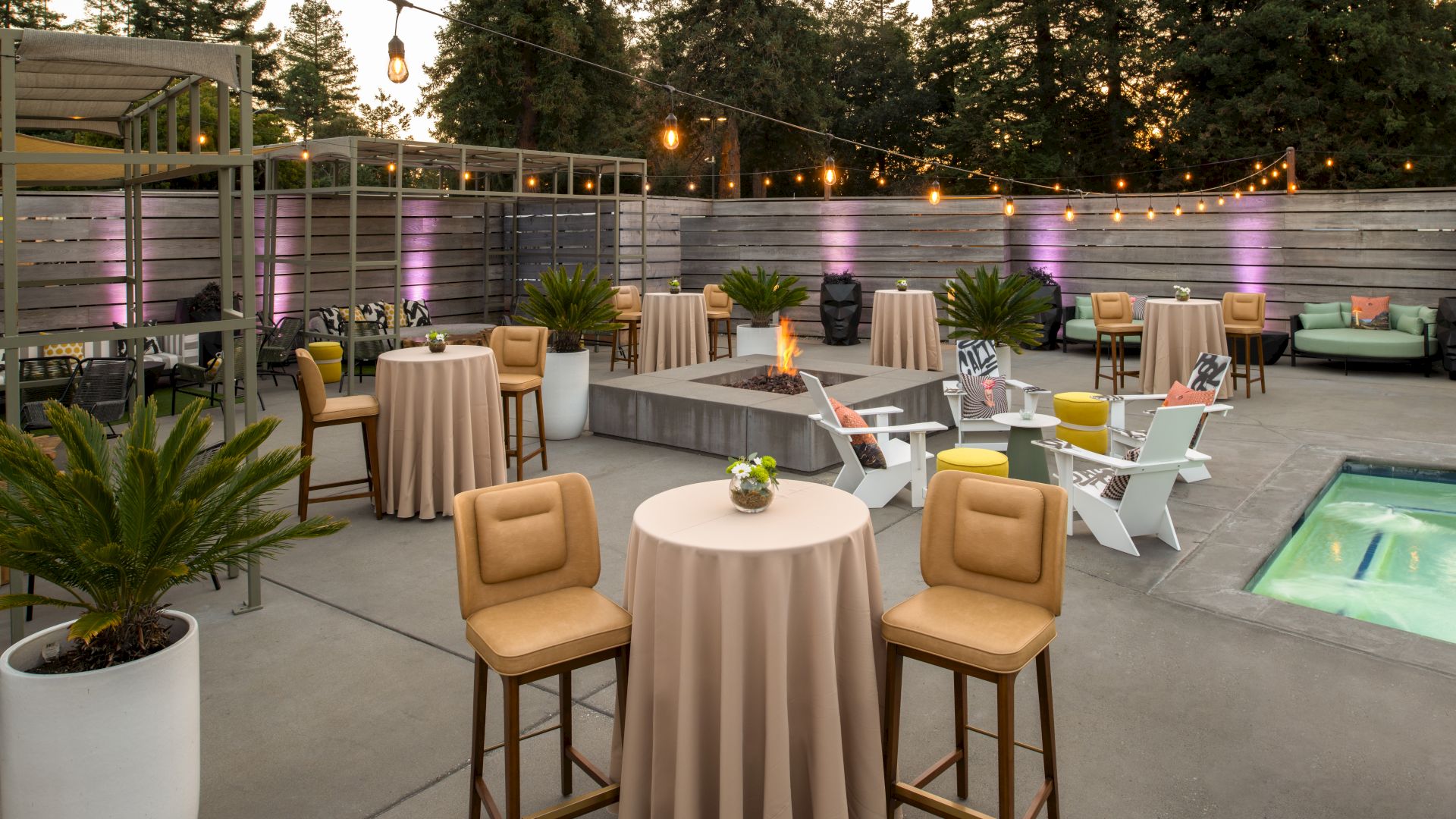 An outdoor patio setup with high tables, chairs, a hot tub, string lights, and surrounding greenery, creating a cozy and inviting ambiance.