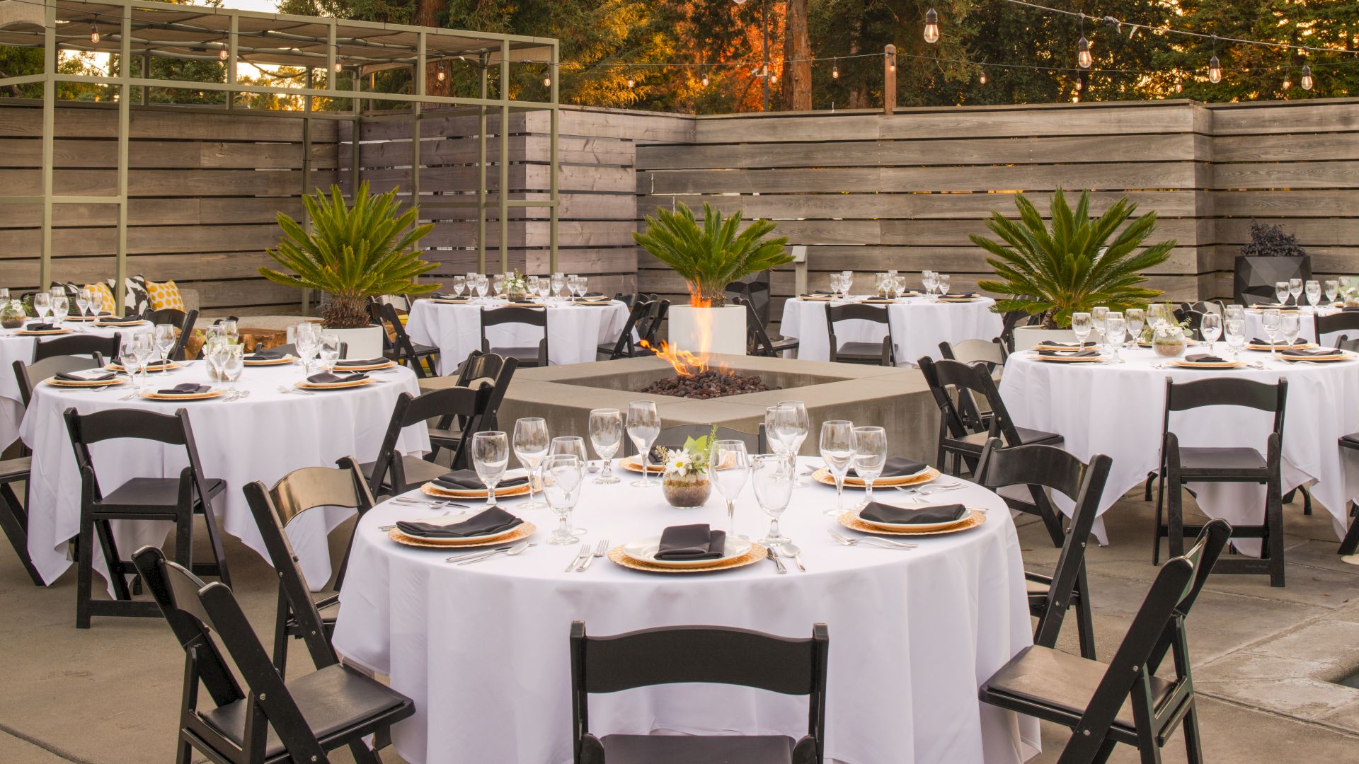 An outdoor event setup with round tables covered in white tablecloths, black chairs, place settings, and a central fire pit surrounded by plants.