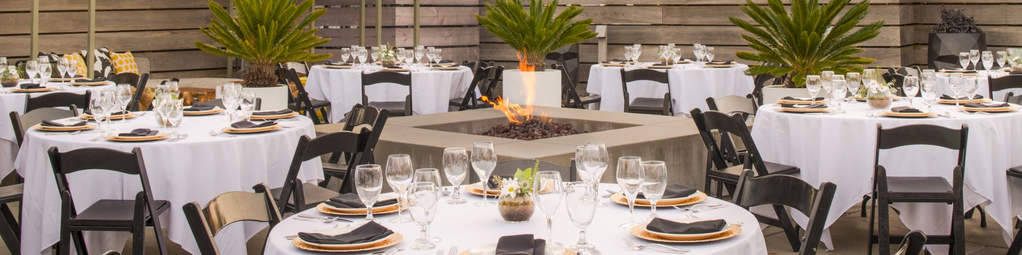 An outdoor event setup with round tables covered in white tablecloths, black chairs, place settings, and a central fire pit surrounded by plants.