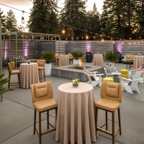 An outdoor patio with tables, chairs, and string lights. There are potted plants, a small pool, and a cozy seating area in the background.