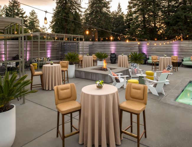 An outdoor patio with tables, chairs, and string lights. There are potted plants, a small pool, and a cozy seating area in the background.