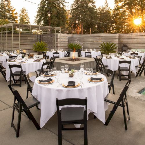 An outdoor event setup with round tables covered in white linens and black chairs, surrounded by trees and a wooden fence, with a small pool nearby.