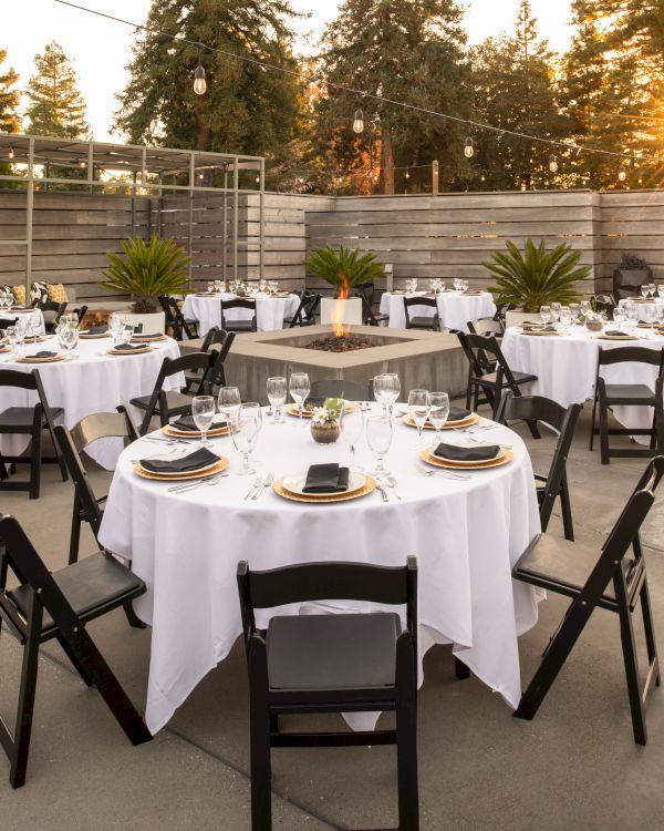 An outdoor event setup with round tables covered in white linens and black chairs, surrounded by trees and a wooden fence, with a small pool nearby.