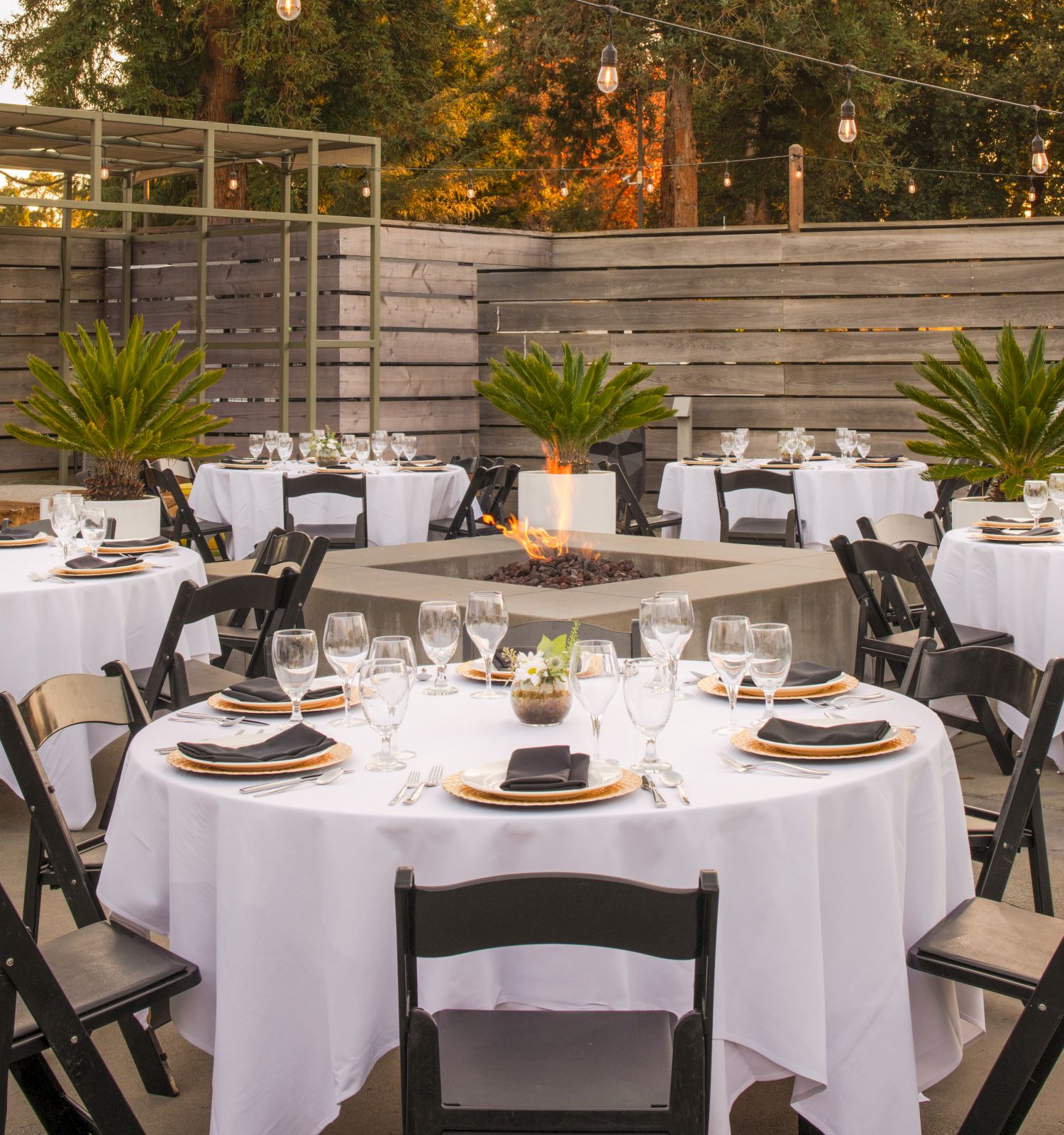 The image shows an outdoor event setup with round tables covered in white tablecloths, black chairs, table settings, and ambient lighting.