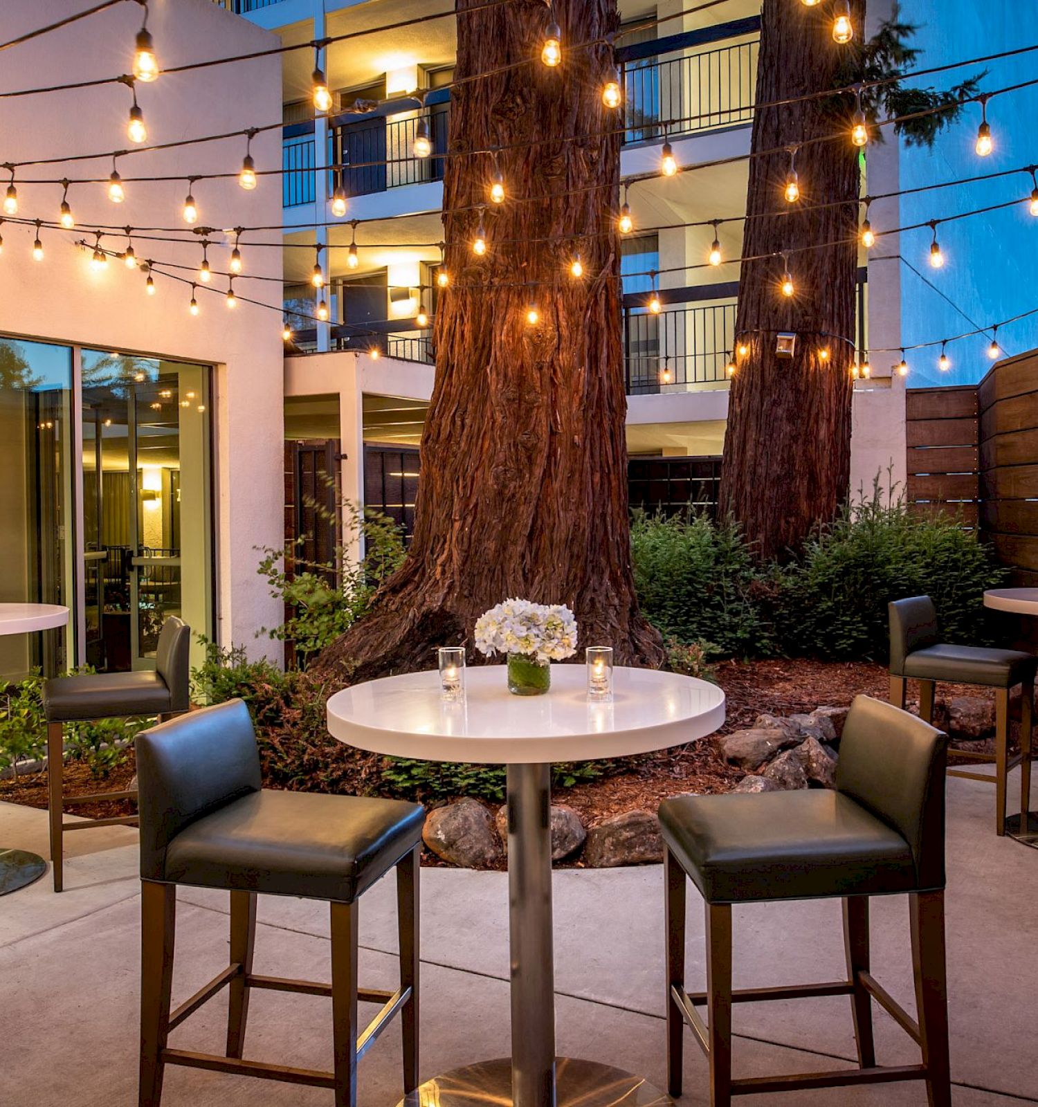 An outdoor seating area with tall tables, chairs, string lights, and floral centerpieces, set against a building and large trees.