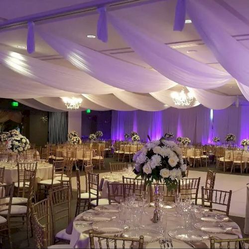 The image shows an elegantly decorated banquet hall with draped ceilings, floral centerpieces, chandeliers, and purple accent lighting.