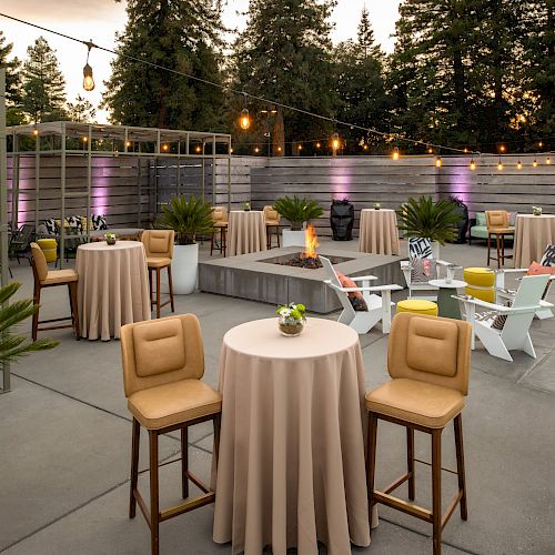 An outdoor event space with round tables, high chairs, plants, string lights, a small pool, and a fire pit area set against a backdrop of trees.