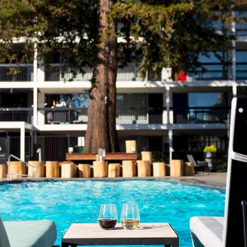 A poolside scene with a table holding two drinks, surrounded by lounge chairs, and a large tree in the background.