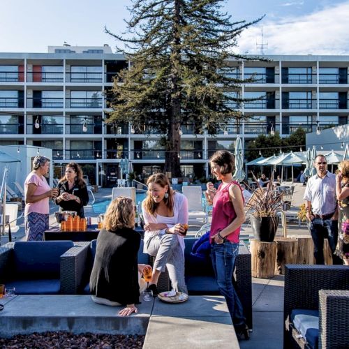 A group of people are gathered outdoors near a pool, socializing and relaxing on lounge furniture with a building in the background.