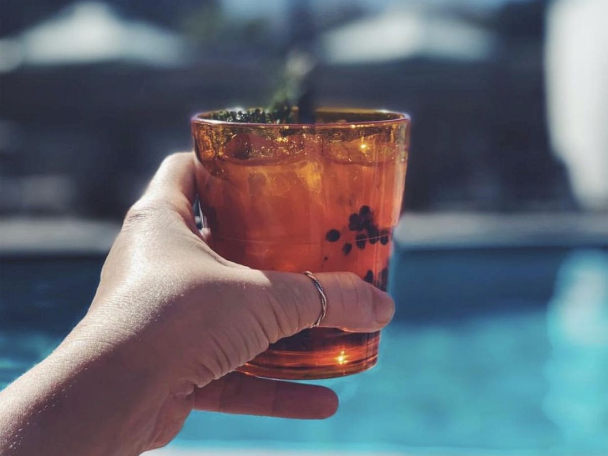 A hand holding a drink in a glass, with a pool in the background. The scene suggests a relaxing time near the pool on a sunny day.