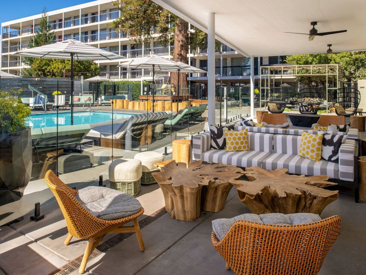 An outdoor seating area by a swimming pool, featuring wicker chairs, wooden tables, patterned cushions, and umbrellas, in a modern setting.