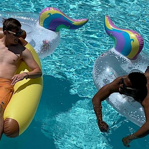 Two people are relaxing on inflatable pool floats in a swimming pool, one on a yellow float and the other on a unicorn-shaped float.