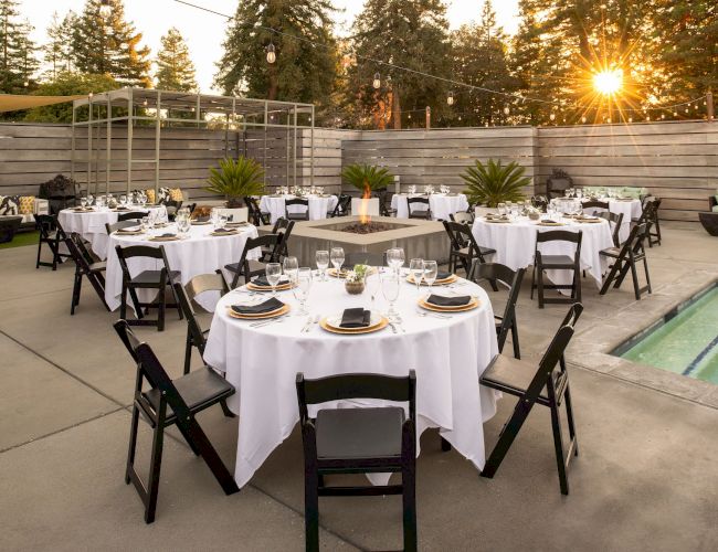 An outdoor dining area set up with round tables, white tablecloths, black chairs, and table settings, surrounded by trees and a setting sun in the background.