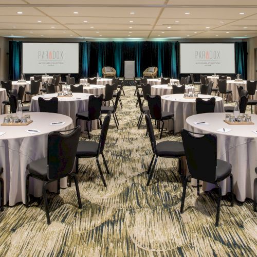 A large conference room set up with round tables and chairs, prepared for a meeting or event. Two screens display the logo for Paradox.