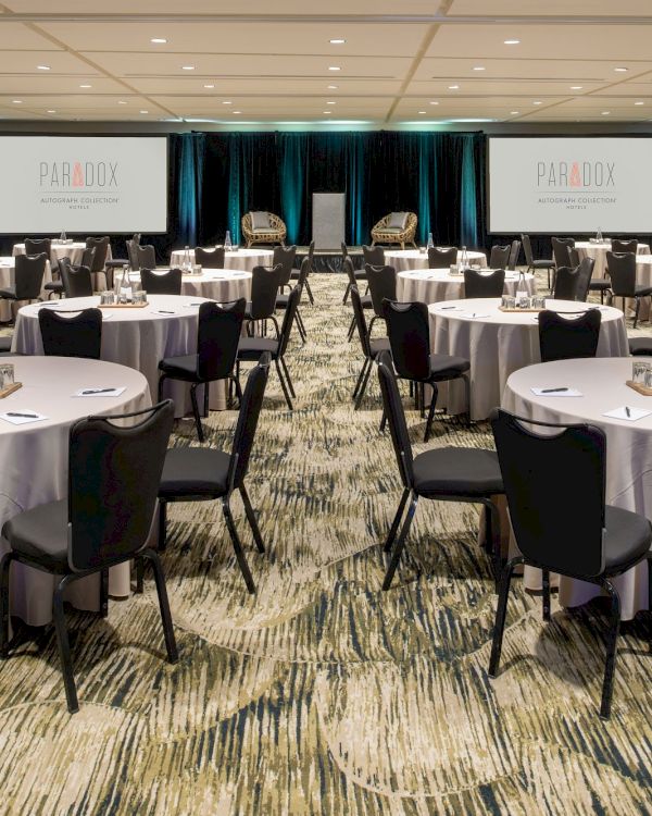 A large conference room set up with round tables and chairs, prepared for a meeting or event. Two screens display the logo for Paradox.