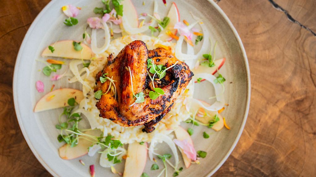 The image shows a dish with grilled chicken atop rice, garnished with microgreens, sliced apples, and thinly sliced onions, on a rustic wooden table.
