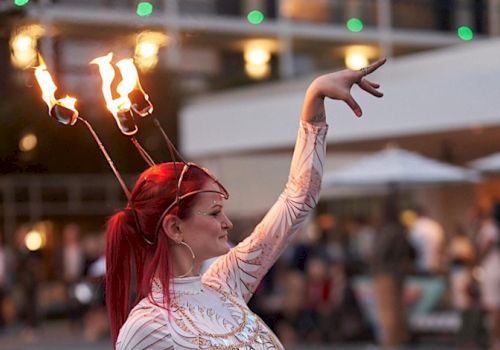 A performer with red hair, wearing an ornate costume, balances lit torches on her head as she poses gracefully outdoors.