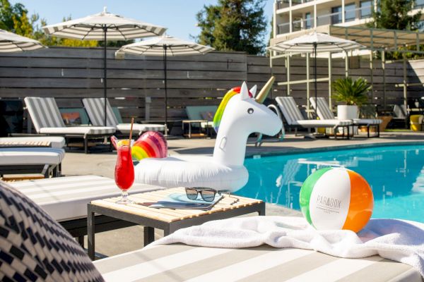 A pool scene with lounge chairs, umbrellas, towels, a unicorn float, a colorful beach ball, and a red drink on a table beside the pool.