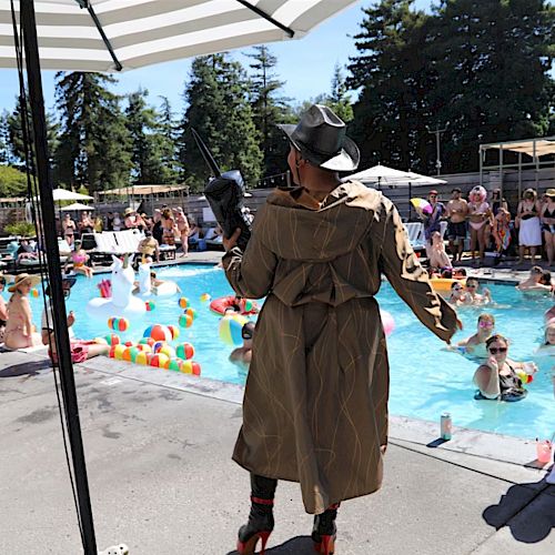 A person in a trench coat and hat entertains people at a lively pool party with floats, drinks, and umbrellas, set in a sunny, forested area.