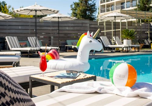 A pool scene with a unicorn float, a cocktail, beach ball, and loungers under umbrellas on a sunny day ends this sentence.
