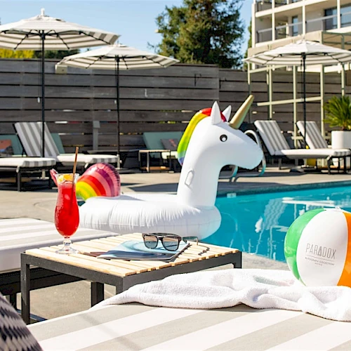 A pool scene with a unicorn float, a cocktail, beach ball, and loungers under umbrellas on a sunny day ends this sentence.