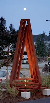 A metal sculpture stands outdoors, surrounded by plants and buildings, under a dusk sky with a visible moon.
