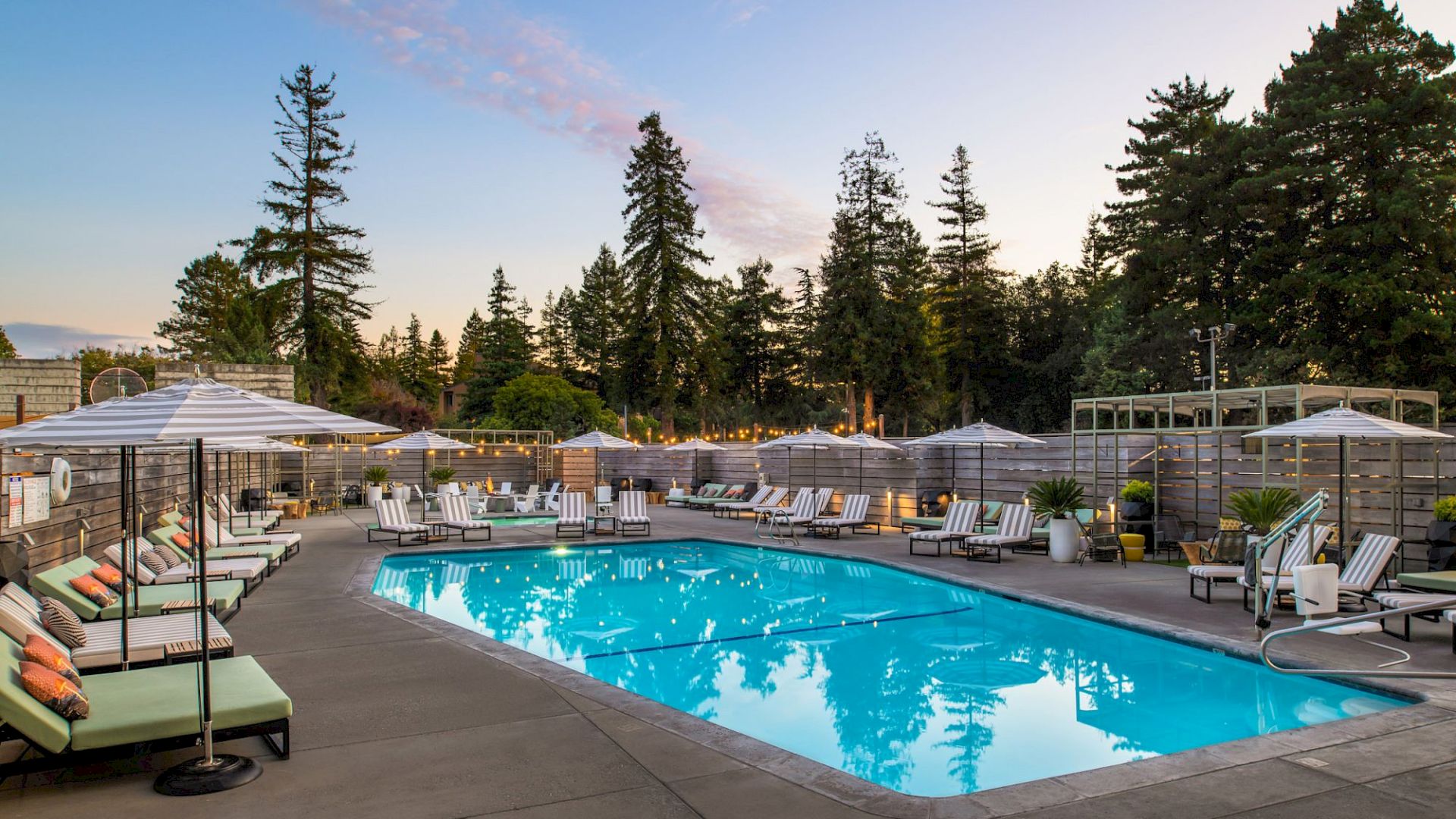 An outdoor swimming pool surrounded by lounge chairs and umbrellas, with tall trees and a blue sky in the background, at what looks like a resort.