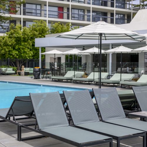 Lounge chairs and umbrellas by a pool, with a modern building in the background and some trees visible.