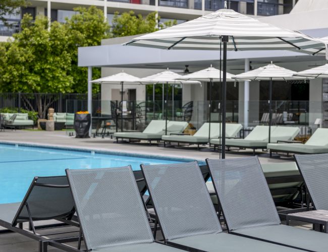 Lounge chairs and umbrellas by a pool, with a modern building in the background and some trees visible.