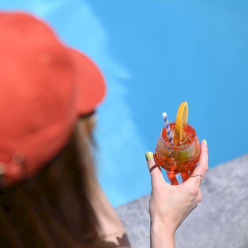 A person in a red cap holds a colorful drink by a poolside, with a clear view of the blue water.