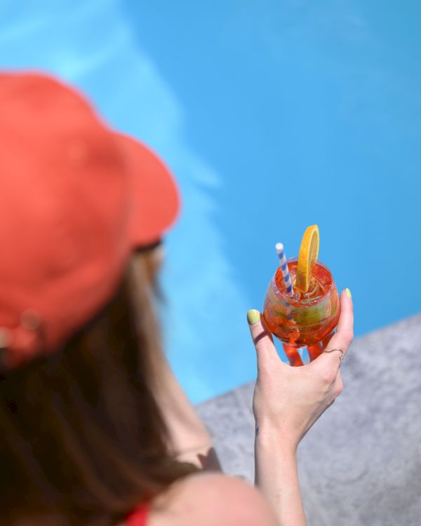 A person in a red cap holds a colorful drink by a poolside, with a clear view of the blue water.