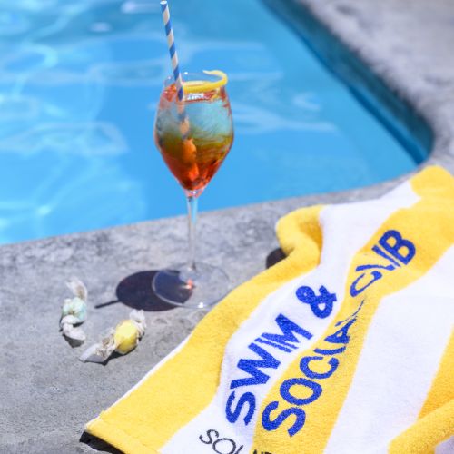 A poolside scene with a cocktail, a yellow towel labeled "Swim & Social Club," and a couple of seashells beside a pool.