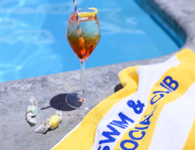 A poolside scene with a cocktail, a yellow towel labeled "Swim & Social Club," and a couple of seashells beside a pool.