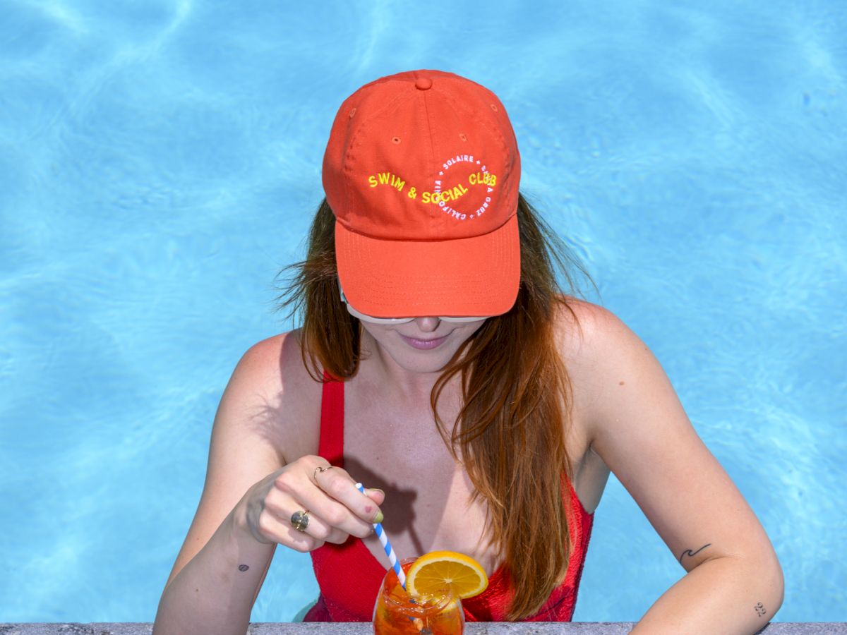 A person in a red swimsuit is in a pool, wearing an orange cap and holding a drink with an orange slice and straw at the edge.