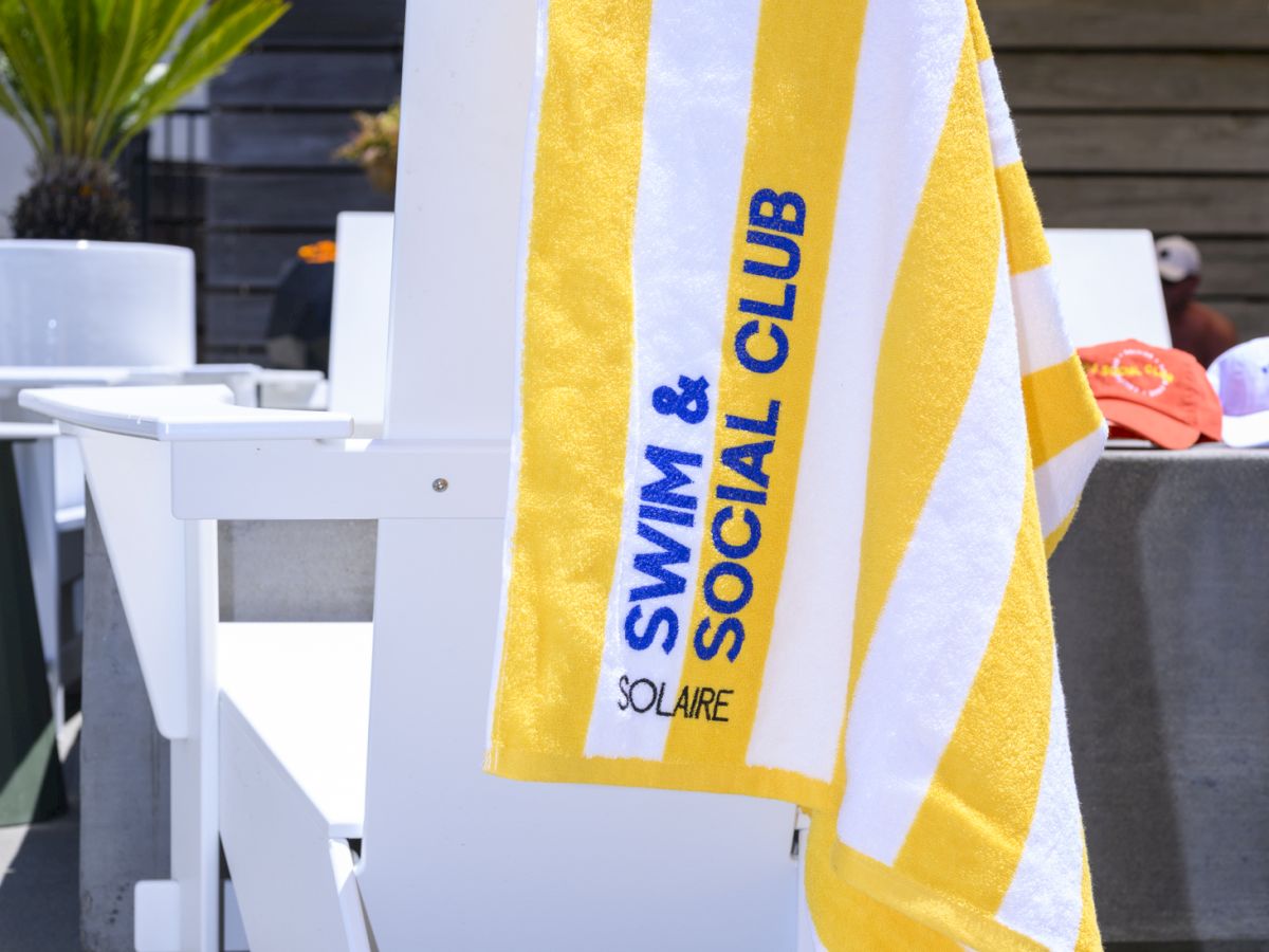 A yellow and white striped towel with "SWIM & SOCIAL CLUB" hangs on a white chair by a poolside area.
