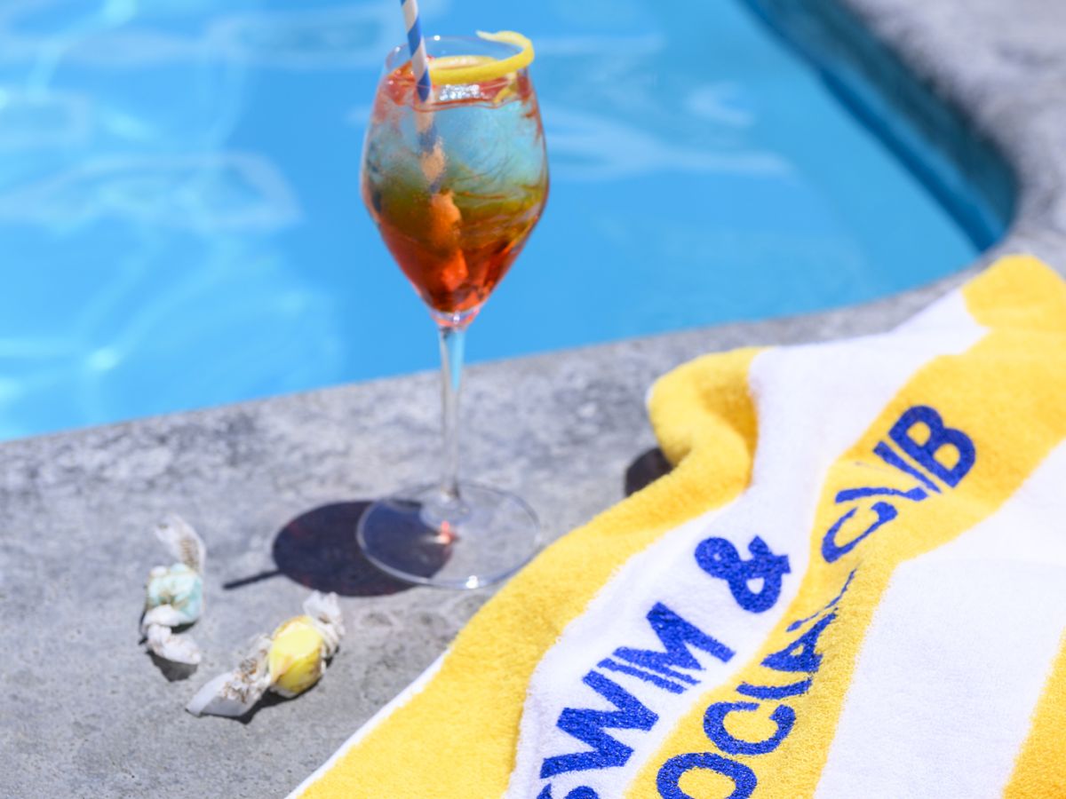 A yellow towel labeled "SWIM & SOCIAL CLUB" by a pool, with a drink in a glass and candy nearby.