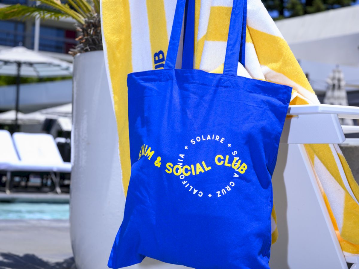 A blue tote bag hangs on a white chair by a yellow-striped towel, next to a large potted plant by a poolside setting.
