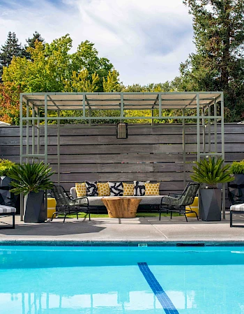 A luxurious pool area with lounge chairs, umbrellas, and a seating area under a pergola. Trees and a building are visible in the background.
